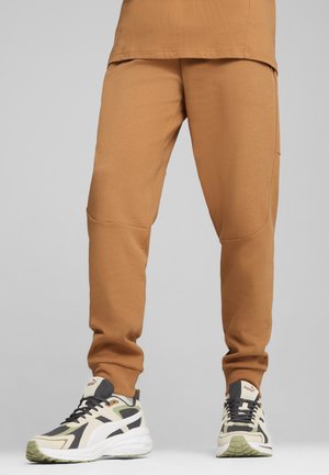 Mand med camel-farvede joggerbukser og hvide, sorte og grønne Puma sneakers, der står imod en ensfarvet baggrund.