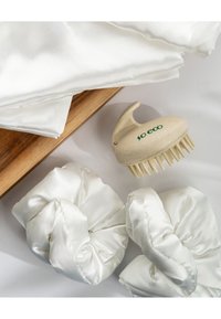 Fascette per capelli in satin bianco, con texture liscia, abbinate a una spazzola per capelli beige con morbide setole in plastica e manico curvo, su una superficie in legno.