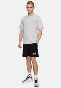 Una camiseta de algodón gris con el texto azul "Fila Sportslife", combinada con pantalones cortos negros que tienen el logo "Fila" en rojo y blanco, y llevada con zapatillas de deporte en blanco y negro.