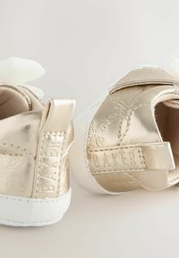 Goldene metallische Babyschuhe mit weißen Sohlen, floralen Prägungen und einem beigen Schleifenakzent an der Vorderseite; Markenetikett an der Ferse.