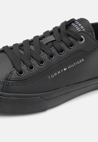 Baskets noires en matière synthétique avec bout rond, lacets plats, et logo brodé "TOMMY HILFIGER" sur le côté. Semelle en caoutchouc texturée.