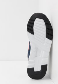 Svart gummisula med vit mellansula, med spår för grepp. Sidan visar blå mocka och grå accenter med röd logotyp.