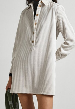 Kobieta w kremowej, prążkowanej sukience z długim rękawem i drewnianymi guzikami, trzymająca w jednej ręce zielono-szary szal.