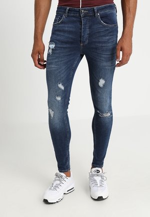 Uomo che indossa jeans skinny blu scuro strappati e sneakers bianche, in piedi davanti a uno sfondo chiaro e semplice.