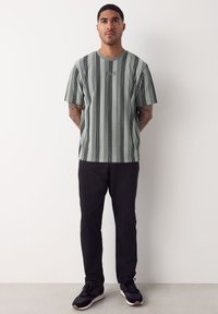 Mann steht mit den Händen hinter dem Rücken, trägt ein grau-schwarz gestreiftes T-Shirt mit vertikalen Streifen, schwarze Hose und schwarze Turnschuhe.