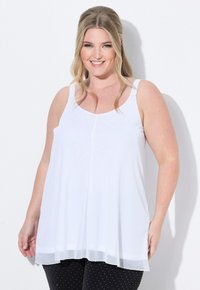 Top senza maniche bianco con vestibilità ampia e orlo trasparente. Presenta bretelle larghe e scollatura rotonda. Indossato con pantaloni neri a pois.