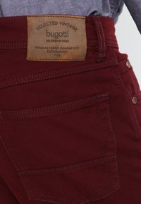 Pantaloni in jeans rossi con etichetta in pelle recante la scritta "bugatti". Caratteristiche includono cuciture multiple e due tasche posteriori con dettagli in ottone.