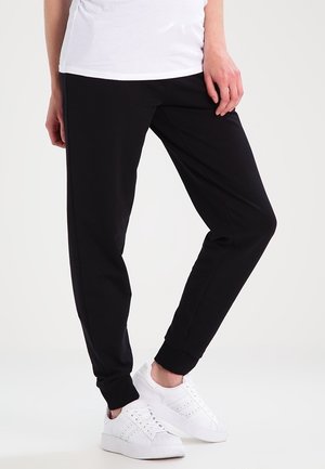 Persona che indossa pantaloni neri a gamba stretta e sneakers bianche, in piedi con una gamba leggermente piegata su uno sfondo chiaro e uniforme.