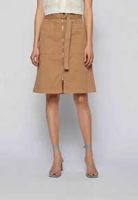 Vaaleanruskea A-linjainen hame, jossa on kaksi etutaskua, etuvetoketju ja kangasnauhavö. Mallilla on jalassa siniset remmillä varustetut korkokengät.