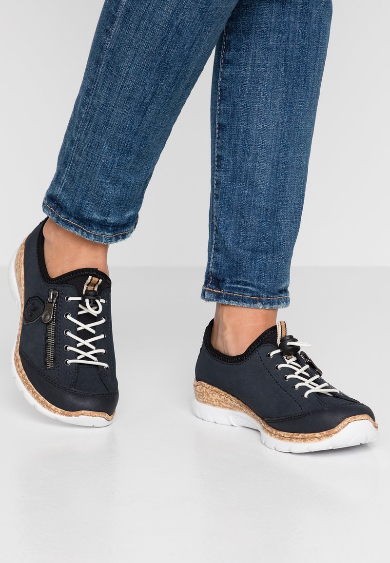 Des baskets noires avec des lacets blancs et une fermeture éclair sur le côté. Accent en liège sur la semelle, tige en tissu texturé, associées à un jean en denim bleu.