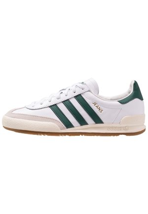 Biały sneaker Adidas z zielonymi paskami, beżowym czubkiem, gumową podeszwą i złotym napisem "JEANS" z boku.