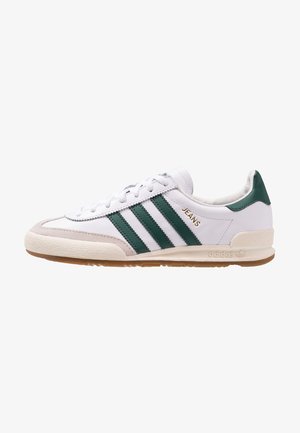 Biały sneaker Adidas z zielonymi paskami, beżowym czubkiem, gumową podeszwą i złotym napisem "JEANS" z boku.