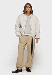 Veste bomber oversized gris clair avec des boutons-pression portée sur un pull gris, associée à un pantalon de survêtement beige fuselé et des chaussures noires à enfiler.