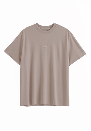 Effen beige T-shirt met korte mouwen en ronde hals, met kleine witte tekst gecentreerd op de borst tegen een witte achtergrond.