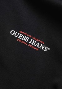 Tessuto nero con testo bianco e rosso che dice "GUESS JEANS" con "AMERICAN TRADITION" sopra e "ORIGINAL DESIGNS" sotto.