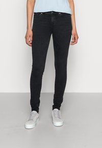 Vaqueros negros ajustados con una textura suave, con una cintura de altura media, cinco bolsillos y herrajes en tono plateado. Combinados con zapatillas gris claro.