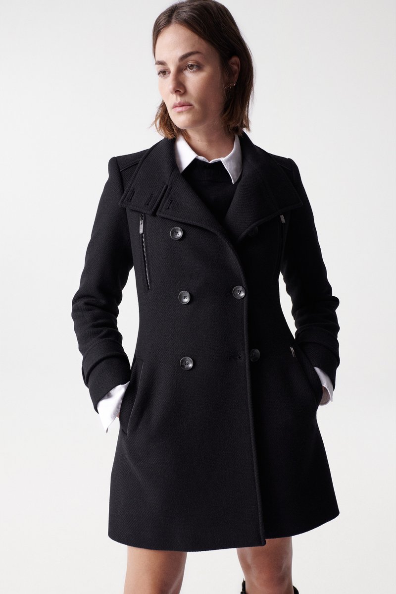 Manteau noir à double boutonnage avec un large col, en tissu texturé, poches latérales et boutons en argent, superposé sur une chemise blanche et un pull.