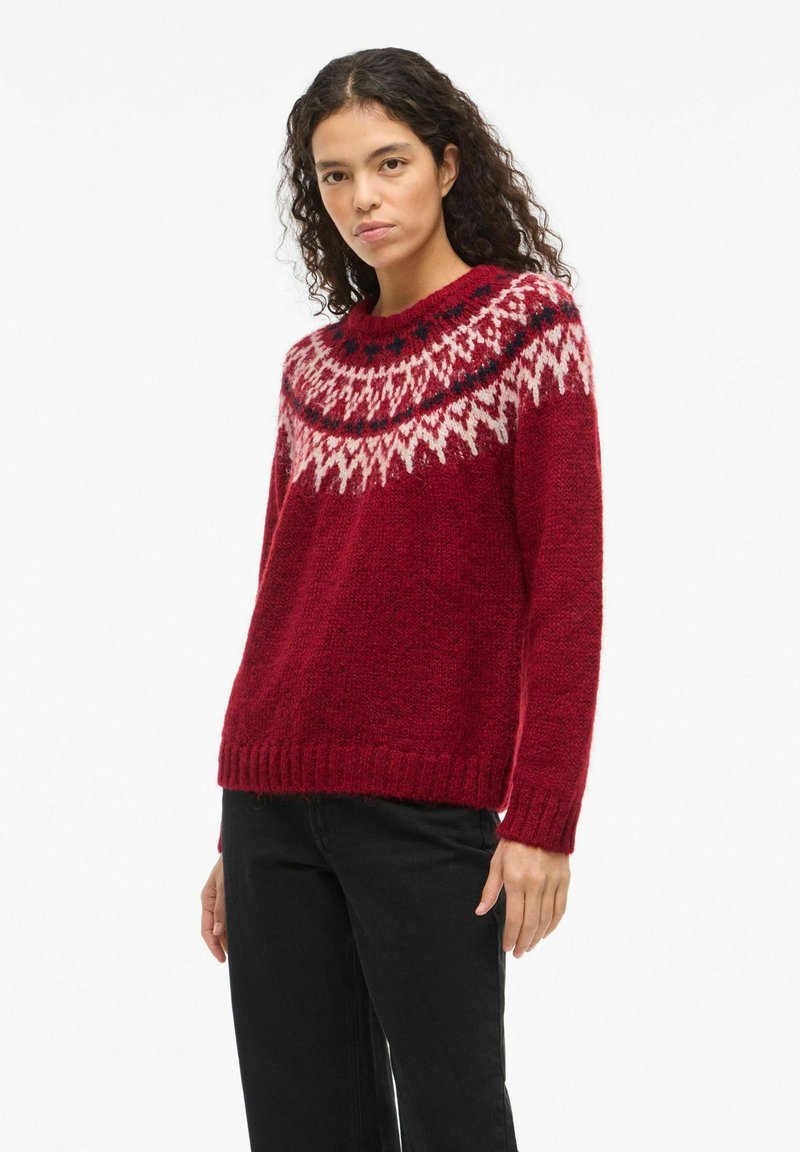 Pull en laine rouge avec un col rond, doté d'un motif géométrique blanc et bleu sur les épaules. Tissu texturé et ourlet côtelé.