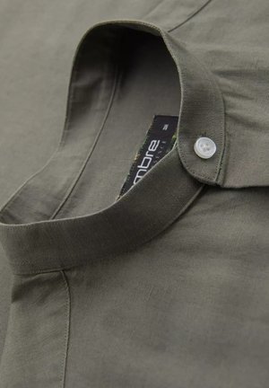 Chemise en lin vert avec un col mandarin, présentant un seul bouton blanc près du cou et une étiquette visible à la couture du col.