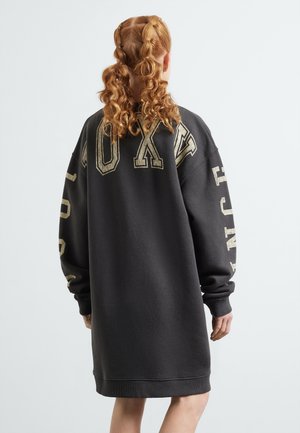 Femme aux cheveux roux bouclés portant une robe sweat-shirt noire surdimensionnée avec de grandes lettres beige au dos et sur les manches, vue de dos.