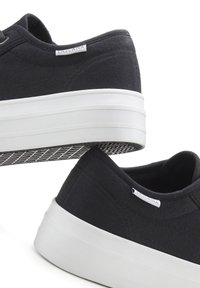 Juodos drobės sportbačiai, turintys storą baltą gumos padą, tekstūruotą raštą ant pado ir mažą logotipo etiketę šone.