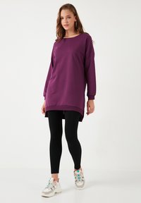Abito felpa oversize viola con maniche lunghe, scollatura rotonda e spacco laterale, abbinato a leggings neri e sneaker chiare.