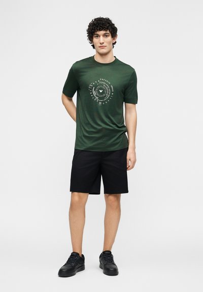 Άνδρας με σγουρά σκούρα μαλλιά, φοράει σκουροπράσινο γραφικό μπλουζάκι, μαύρο σορτς και μαύρα αθλητικά παπούτσια, στέκεται μπροστά σε ένα απλό φόντο.