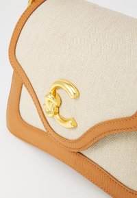 Borsa in tela beige con bordo in pelle marrone chiaro, caratterizzata da un accento in metallo dorato con logo e texture liscia. Bordo curvo e chiusura a patta.