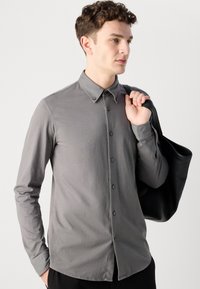 Grijze longsleeve overhemd met knoopsluiting en een puntige kraag, met een subtiele glans, gecombineerd met een zwarte tote bag. Het overhemd heeft een getailleerde pasvorm.