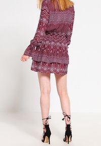 Maronfarvet kjole med hvide mønstre, flagrende ærmer og lagdelte flæser i kanten. Sammen med sorte strop sandaler. Vist fra ryggen.