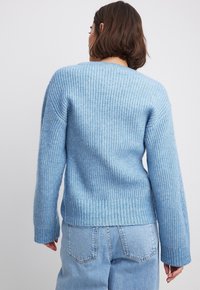 Modrý hrubý pletený svetr s žebrovanou strukturou, širokými rukávy a rovnou spodní částí. Zadní strana má vysok�ý límec a otevřené detaily předení.