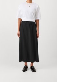 Svart, knälång kjol med en slät textur och en rynkad elastisk midja, tillsammans med en löst sittande vit T-shirt och svarta slip-on skor.