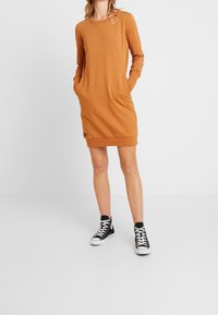 Robe orange à manches longues avec un col rond, des poches latérales et des détails de boutons sur l'épaule. Associée à des baskets montantes noires.