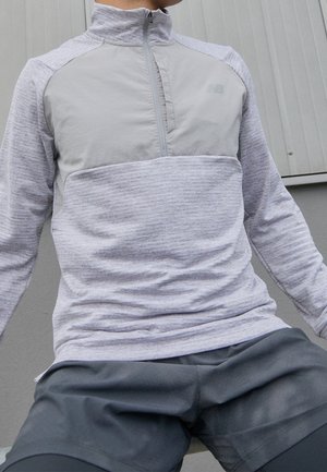 Person trägt einen hellgrauen Pullover mit Viertel-Reißverschluss und dunkelgraue Sportshorts, sitzt vor einem grauen Wandhintergrund.