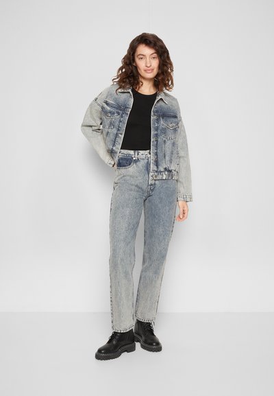 Casaco de denim em azul desbotado com grandes bolsos e botões pretos, combinado com uma blusa preta e jeans azul claro, finalizado com botas pretas.