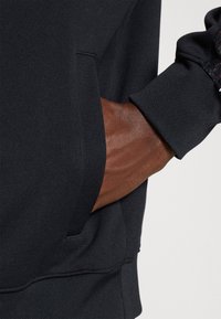 Sudadera negra con un bolsillo y puños texturizados. El material parece suave y elástico, mostrando un primer plano de una mano descansando dentro del bolsillo.