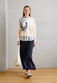 Sudadera en crema con un gráfico de ave, superpuesta sobre una blusa de rayas, combinada con pantalones anchos oscuros y un bolso beige claro.