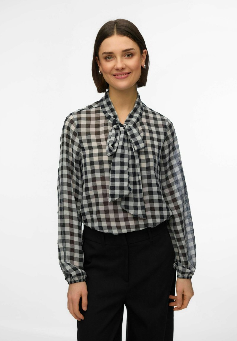 Femme aux cheveux bruns courts portant un chemisier à carreaux noir et blanc avec un nœud noué au col et un pantalon noir, souriant doucement.