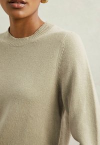 Šviesiai beige megztinis su apvaliu kaklu ir raukiniais ant rankovių. Medžiaga yra minkšta ir lygi, su subtiliomis siuvimo detalėmis ant pečių.