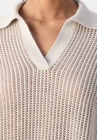 Gros plan sur une personne portant un pull beige en maille ajourée à encolure en V avec un col souple, laissant la peau visible sous le tissu.