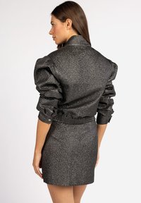 Schwarze, strukturierte, kurz geschnittene Jacke mit puffigen Ärmeln und elastischem Taillenbund, kombiniert mit einem passenden, figurbetonten Rock. Glatte und reflektierende Oberfläche.