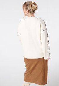 Vrouw met blond haar in een bun, gekleed in een crèmekleurige trui met zwarte trim en een bruine suède rok, staande tegen een witte achtergrond.