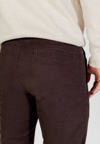 Pantaloni marroni in velluto di cotone con una texture liscia, dotati di tasche posteriori e un design aderente, abbinati a un maglione crema chiaro.
