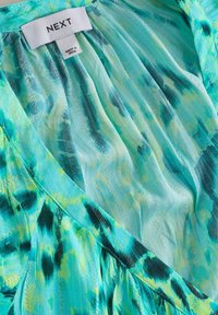 Gros plan d'un tissu à motifs bleu-vert avec une étiquette de vêtement blanche cousue sur laquelle est inscrit "NEXT" et "Made in India."