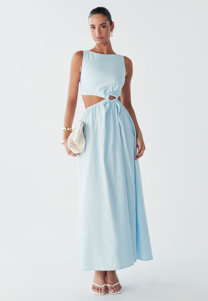 Le modèle pose dans une robe maxi bleu clair à découpes avec une taille nouée, tenant un sac crème et portant des sandales blanches à talons.