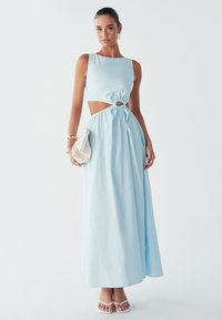 Iškirptų detalių ilga suknele šviesiai mėlynos spalvos audinyje, su prigludusiu liemeniu, susuktos priekinės dalies akcentu ir laisvai krentančia suknele. Porą baltų basučių su dirželiais.