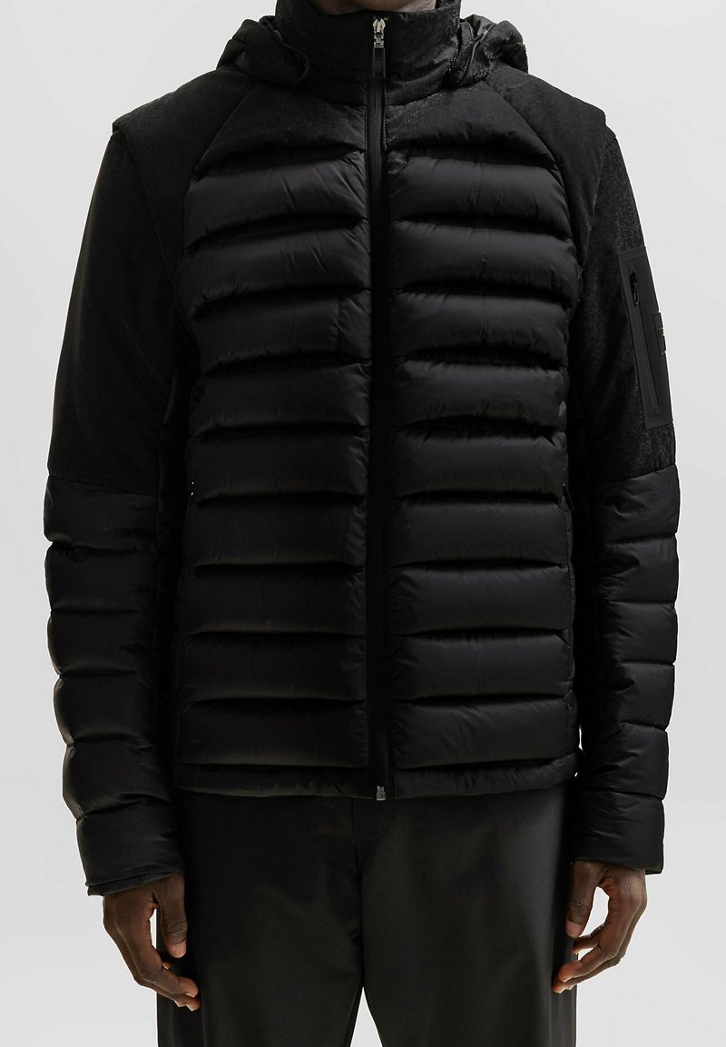 Homme portant une doudoune noire à capuche avec des sections rembourrées horizontales et une fermeture éclair devant, sur un fond uni.