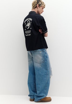 Młoda osoba z krótkimi blond włosami, ubrana w czarną koszulkę z motywem skorpiona oraz luźne niebieskie dżinsy na jednolitym białym tle.