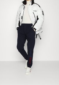 White puffer jacket with black accents, featuring a logo. Navy sweatpants with red and blue logo text along the side. White sneakers.