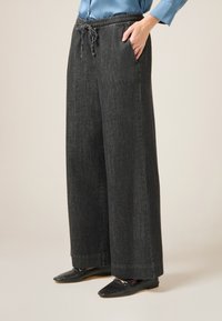 Weite schwarze Denim-Hosen mit elastischem Bund, Kordelzugdetails und Seitentaschen; kombiniert mit flachen schwarzen Schuhen.